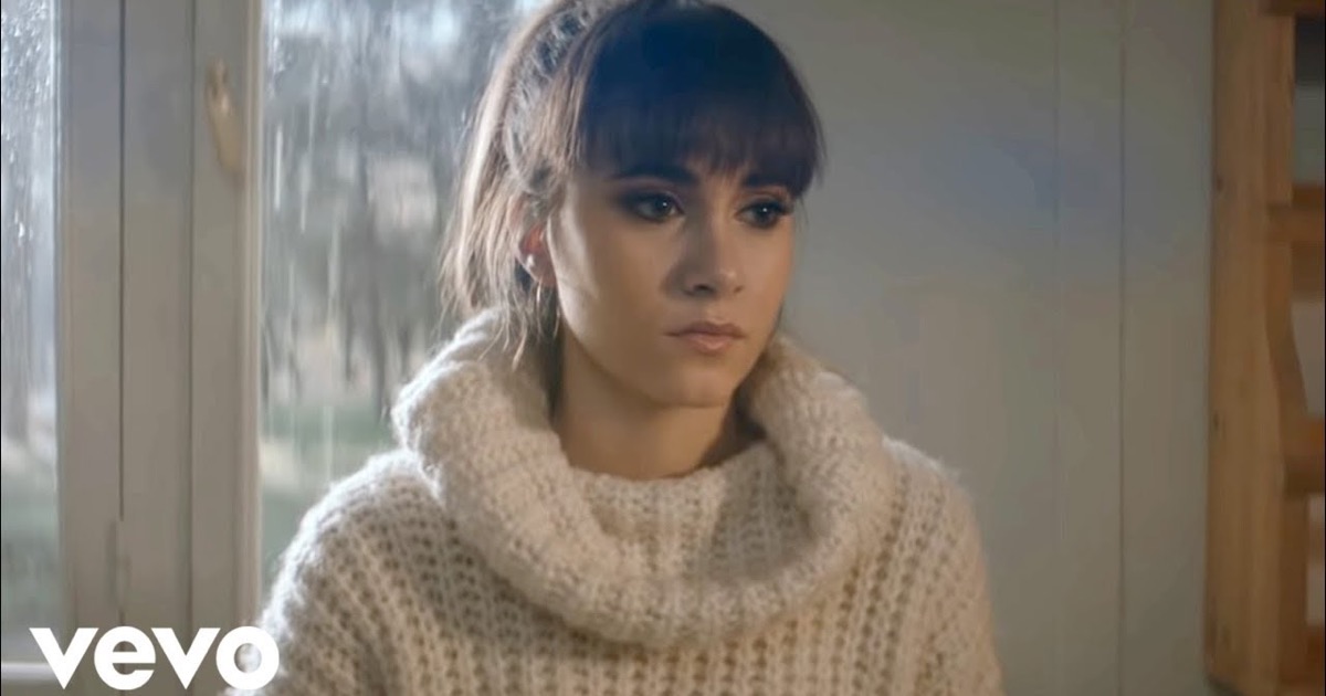 Aitana en el videoclip oficial de Vas A Quedarte (2018): primer plano de la cantante con iluminación cálida, uno de sus temas más reproducidos en YouTube