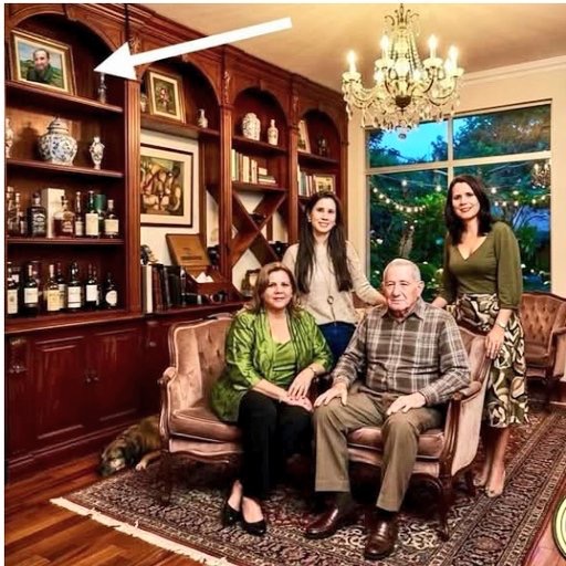 Salón con Raúl Castro sentado junto a mujeres de la familia Rueda Cardero; flecha blanca señala retrato enmarcado de Alejandro Castro Espín en la estantería